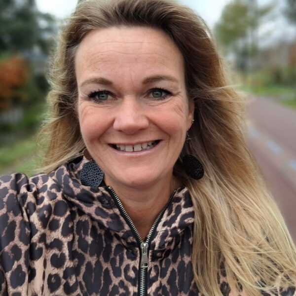 Persoon in een jas met dierenprint staat op een fietspad en kijkt richting de camera, met bomen en groen in de onscherpe achtergrond.
