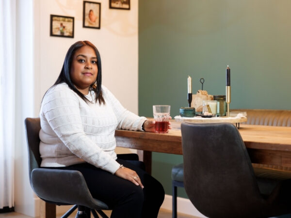 Persoon in een witte trui zit aan een houten eettafel met een glas rode drank. Op de tafel staan kandelaars en decoratieve voorwerpen.