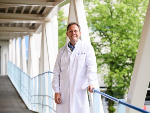 Persoon in een witte laboratoriumjas staat op een loopbrug en houdt een leuning vast. Op de achtergrond zijn bomen en een gebouw zichtbaar.