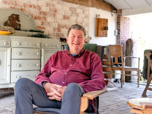 Persoon zit in een houten stoel op een overdekt terras met bakstenen muur en landelijk interieur op de achtergrond.