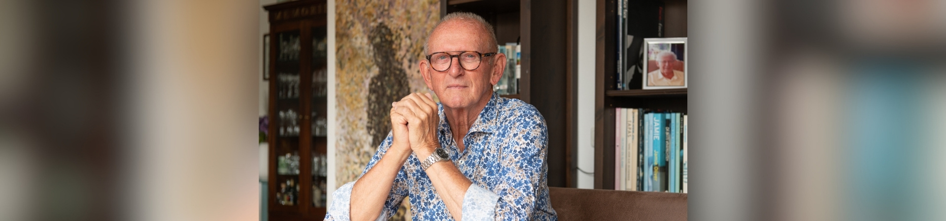 Portret van een persoon aan een houten tafel in een woonruimte met boekenkast op de achtergrond.