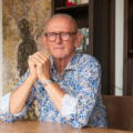 Portret van een persoon aan een houten tafel in een woonruimte met boekenkast op de achtergrond.