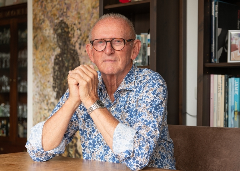 Portret van een persoon aan een houten tafel in een woonruimte met boekenkast op de achtergrond.