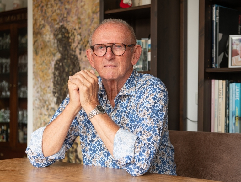 Portret van een persoon aan een houten tafel in een woonruimte met boekenkast op de achtergrond.